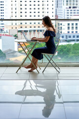 Business remotely. A girl works at her laptop on a balcony in the city center with a landscape view