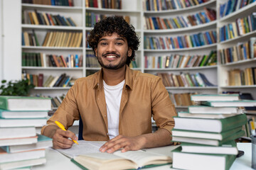 Happy and smiling student studying inside academic library, man smiling and looking at camera doing...