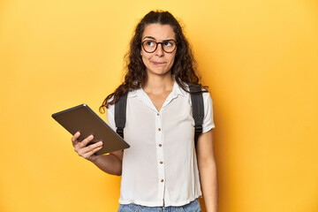 Student holding a tablet, glasses, backpack on, confused, feels doubtful and unsure.