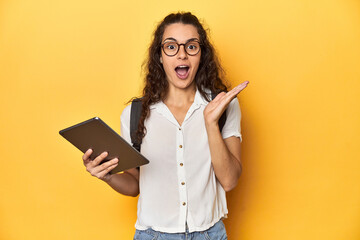 Student holding a tablet, glasses, backpack on, surprised and shocked.