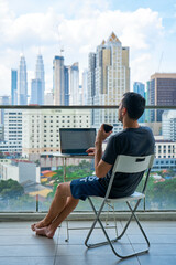 Business remotely. A man works at him laptop on a balcony in the city center with a landscape view.
