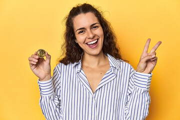 Woman holding a physical Bitcoin coin, joyful and carefree showing a peace symbol with fingers.