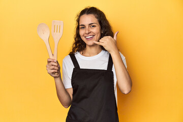 Woman with apron, wooden cooking utensils, yellow, showing a mobile phone call gesture with fingers.
