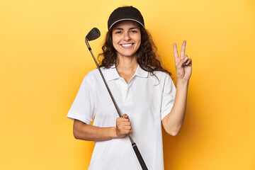 Golfer woman with cap, golf polo, yellow studio, showing number two with fingers.