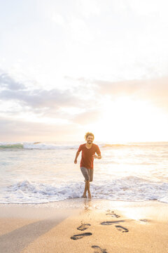 Vertical image of a smiling young man running through the waves at the shore of the beach on a summer day with beautiful sunset light.