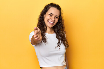 Young Caucasian woman, yellow studio background, stretching hand at camera in greeting gesture. © Asier