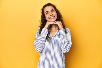 Young Caucasian woman, yellow studio background, keeps hands under chin, is looking happily aside.