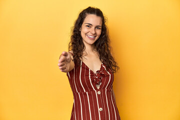 Young Caucasian woman, yellow studio background, stretching hand at camera in greeting gesture.
