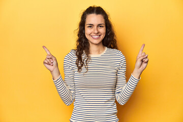 Young Caucasian woman, yellow studio background, indicates with both fore fingers up showing a...