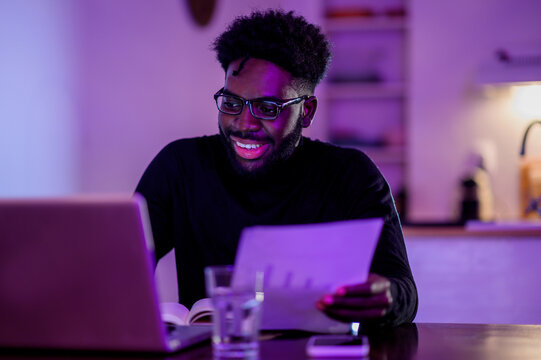 A happy african american casual businessman is sitting at home at night and working on a project on a laptop.