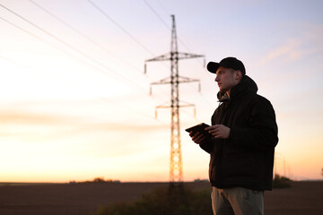 Young power engineer holds digital tablet against high voltage tower at field. Electrical engineer...