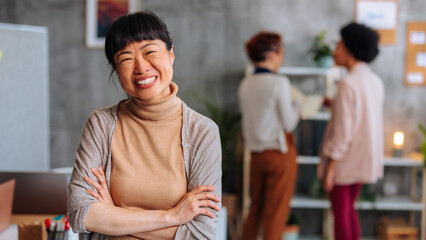 Joyful Asian business woman smiling with arms crossed while standing in busy startup office.