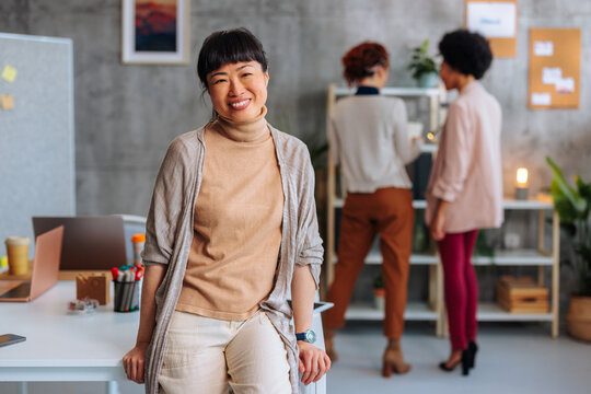 Young Asian Business Woman Smiling While Sitting On Desk In Company Office.