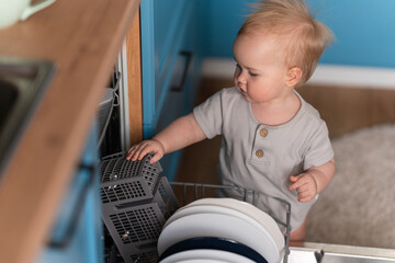 Toddler baby helps to get plates out of the dishwasher at home in the kitchen