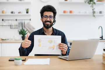 Portrait of indian male in headset and glasses posing with infographics on paper at desk with digital devices. Smiling entrepreneur presenting marketing report on online meeting from home workplace.
