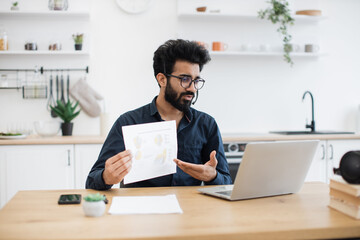 Fototapeta premium Confident mature adult with wireless headset pointing at graphic materials while giving online presentation from home. Focused indian worker providing statistical analysis using modern technologies.
