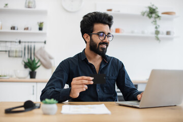 Attractive bearded man in spectacles holding blank credit card while typing on portable computer in kitchen interior. Happy arabian gentleman doing online banking transaction while working remotely.