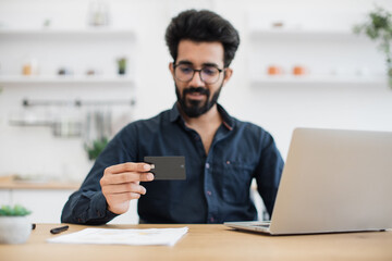 Fototapeta na wymiar Focus on young man smiling at blank debit card while sitting at writing desk with computer in kitchen of apartment. Delighted indian male making reservation on e-commerce website without leaving home.