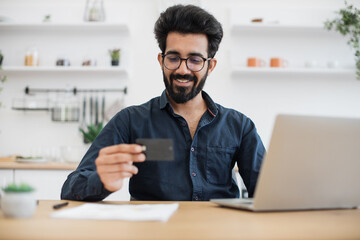 Focus on young man smiling at blank debit card while sitting at writing desk with computer in kitchen of apartment. Delighted indian male making reservation on e-commerce website without leaving home.