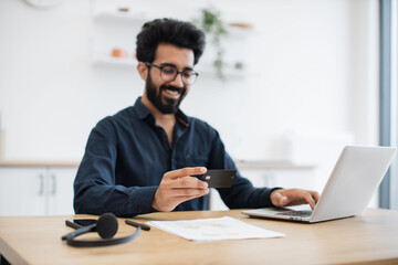 Fototapeta premium Joyful arabian man in casual clothes using black credit card while running portable computer in workplace. Smiling bearded person in glasses enjoying virtual shopping using online wallet from home.