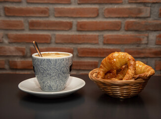 Detail shot of a breakfast with a delicious and shiny croissant in a wicker basket accompanied by a cup of coffee with milk on a plain black table with a brick wall in the background.