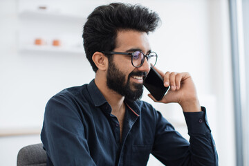 Close up portrait of handsome arabian adult wearing glasses and button-down shirt being involved in phone discussion. Joyous chief manager negotiating contract being outside of corporate offices.