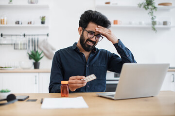 Fototapeta premium Feverish bearded man in casual clothes touching forehead while looking at laptop camera in dining room of apartment. Unwell arabian manager arranging virtual appointment with health professional.