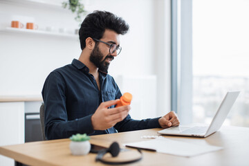 Handsome mature person in spectacles holding bottle of pills while sitting in home office with laptop at midday. Indian businessman examining medicine prescribed after online consultation by doctor.