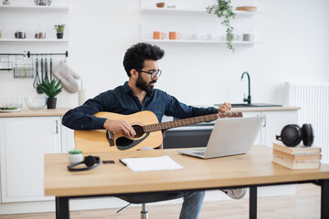 Young male musician in dark shirt tuning acoustic guitar using laptop on writing desk in modern...