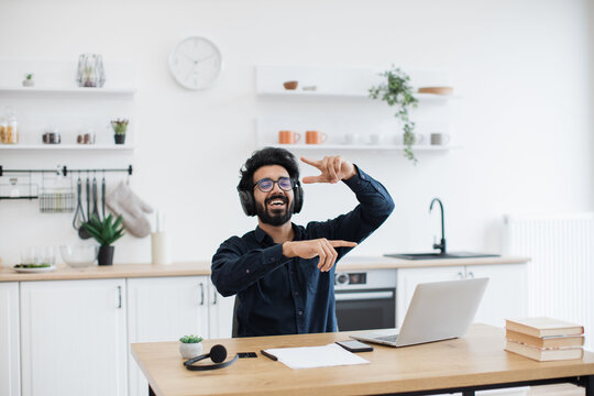 Emotional mature adult with raised fists listening to favourite songs while resting at wooden desk in kitchen interior. Indian music enthusiast rocking out to energetic tunes in black headphones.