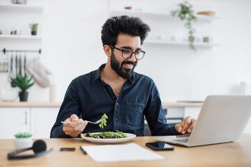 Smiling mature man in business casual clothes eating healthy salad while looking at laptop screen...