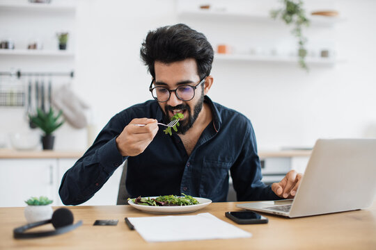 Young indian businessman tasting fresh greens in vegan meal while sitting at office desk in bright apartment. Self-employed worker enjoying nutritious food during lunch hour while telecommuting.
