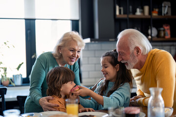 Two playful smiling kids boy and girl having donuts for dessert after breakfast with caring grandparents beautiful senior woman and handsome senior man. Sister sharing delicious donut with brother.