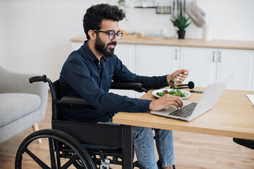 Positive indian adult with disability having meal while scrolling webpages on laptop in spacious dining room. Well-organised employee combining lunch break and flexible schedule at distance work.