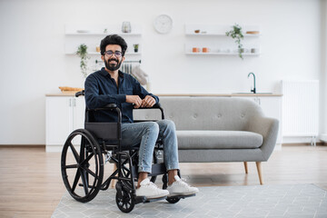 Full length portrait of arabian wheelchair user smiling at camera while resting in open-plan...