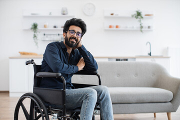 Fototapeta na wymiar Portrait of smiling indian man with disability wearing glasses and casual outfit posing in spacious dining room. Positive young adult feeling optimistic while making recovery from injury at home.