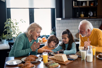 Happy seniors having breakfast at kitchen table with grandkids at home, looking at them with love and kindness. Smiling senior woman and senior man enjoying their time with kids at breakfast. - obrazy, fototapety, plakaty