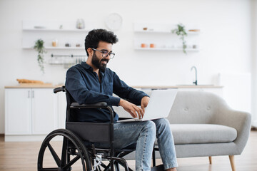 Joyful indian man in wheelchair holding laptop on knees while staying in middle of bright open-plan kitchen. Happy young adult surfing internet webpages while recovering after accident at home.