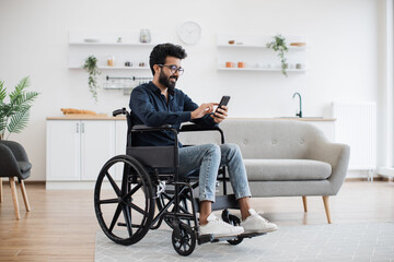Full length view of indian male with disability using smartphone while resting in modern kitchen of...
