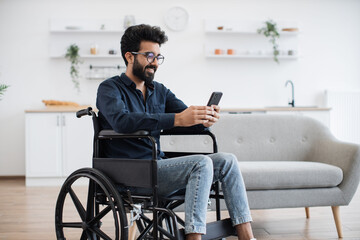 Smiling arabian adult in wheelchair looking at cell phone screen while having rest in comfortable dining room. Cheerful positive man checking social media using internet connection in apartment. - obrazy, fototapety, plakaty