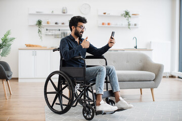 Cheerful wheelchair user in spectacles and casual outfit utilizing phone webcam while enjoying free time indoors. Bearded indian male receiving online conference call via mobile application about job.