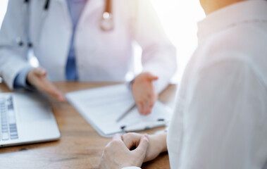 Doctor and patient sitting and discussing something at wooden table while using a laptop computer. Focus is on patient's hands. Medicine concept