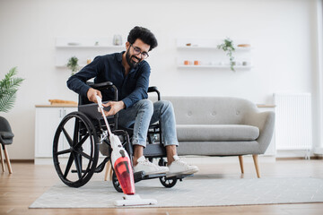 Fototapeta na wymiar Relaxed adult person in jeans taking lightweight stick vacuum while using wheelchair in spacious flat. Cheerful indian homeowner enjoying housework with modern appliances while waiting for guests.