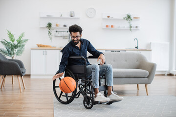 Cheerful bespectacled adult with mobility impairment dribbling ball in studio apartment on weekend. Happy indian in jeans and dark shirt staying active with disability while playing game for pleasure.