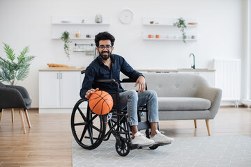 Cheerful bespectacled adult with mobility impairment dribbling ball in studio apartment on weekend....