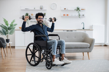 Full length view of handsome indian man in wheelchair lifting weights while exercising with limited mobility indoors. Smiling adult using moderate-intensity activity during home workout in kitchen.