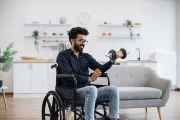 Delighted arabian male working out with dumbbell while staying in good mood on sunny day at home. Smiling mature person in spectacles increasing endurance while doing exercise with disability.