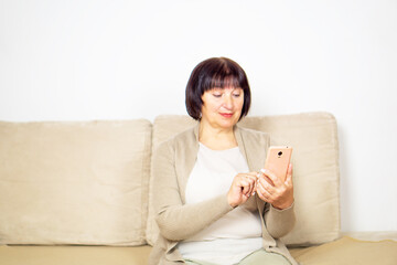 Happy relaxed older woman sitting on comfortable sofa at home and using a mobile phone.