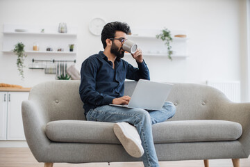 Cheerful indian mature person smiling at portable computer screen while drinking hot coffee in open-plan kitchen. Stylish bespectacled man blogging social media site while enjoying pastime at home.