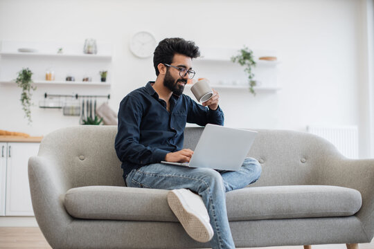 Handsome Bearded Man With Cup Of Tea In Hand Using Modern Laptop While Resting On Sofa At Home. Young Arabian Person In Glasses Surfing Internet Webpages At Leisure Time Indoors.